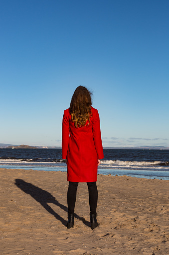 Woman wearing red coat on the beach, looking out to sea