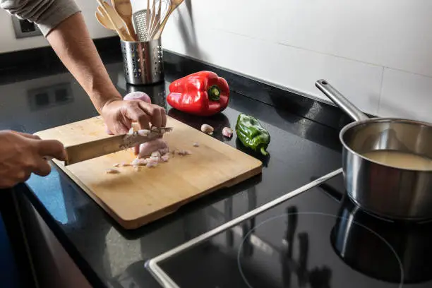 male hands chopping onions on a wooden board with a chef's knife. He has vegetables and kitchen utensils needed for his recipe