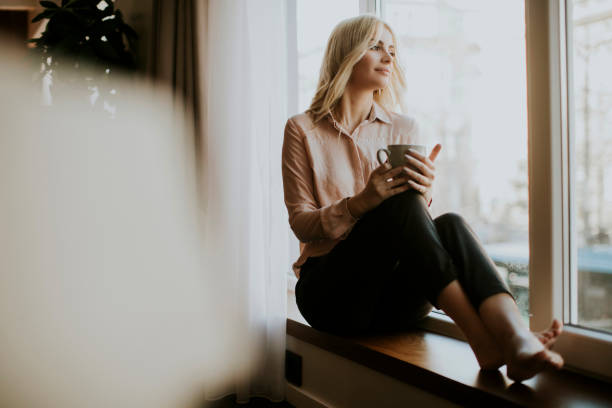 istock Blonde young woman drinking coffee by window 949115272