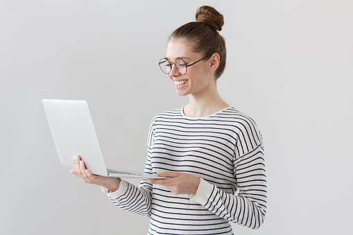 Shot of beautiful young girl standing isolated against gray background wearing big round glasses, standing and looking at screen of laptop that she holds, smiling happily at positive news from web.