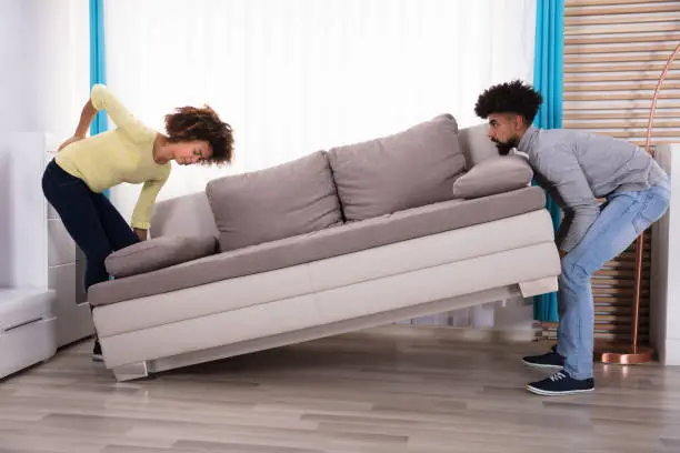 Woman Having Backpain While Lifting The Sofa With Her Husband In Living Room
