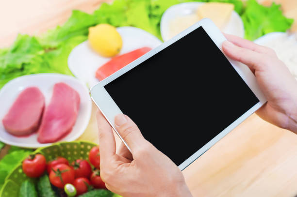 istock Close-up top view of female hands holding a tablet computer with a blank display surrounded by healthy food on a home kitchen table. Workpiece to insert the design of the mobile application into the screen. Products of Greek cuisine raw seafood and fresh 949187678