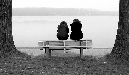 Two girls sitting on the bench between  two trees.