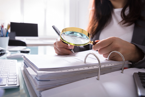 Close-up Of A Businesswoman's Hand Checking Bill Through Magnifying Glass At Workplace