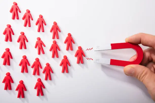 Close-up Of A Human Hand Attracting Red Human Figures With Horseshoe Magnet On White Background