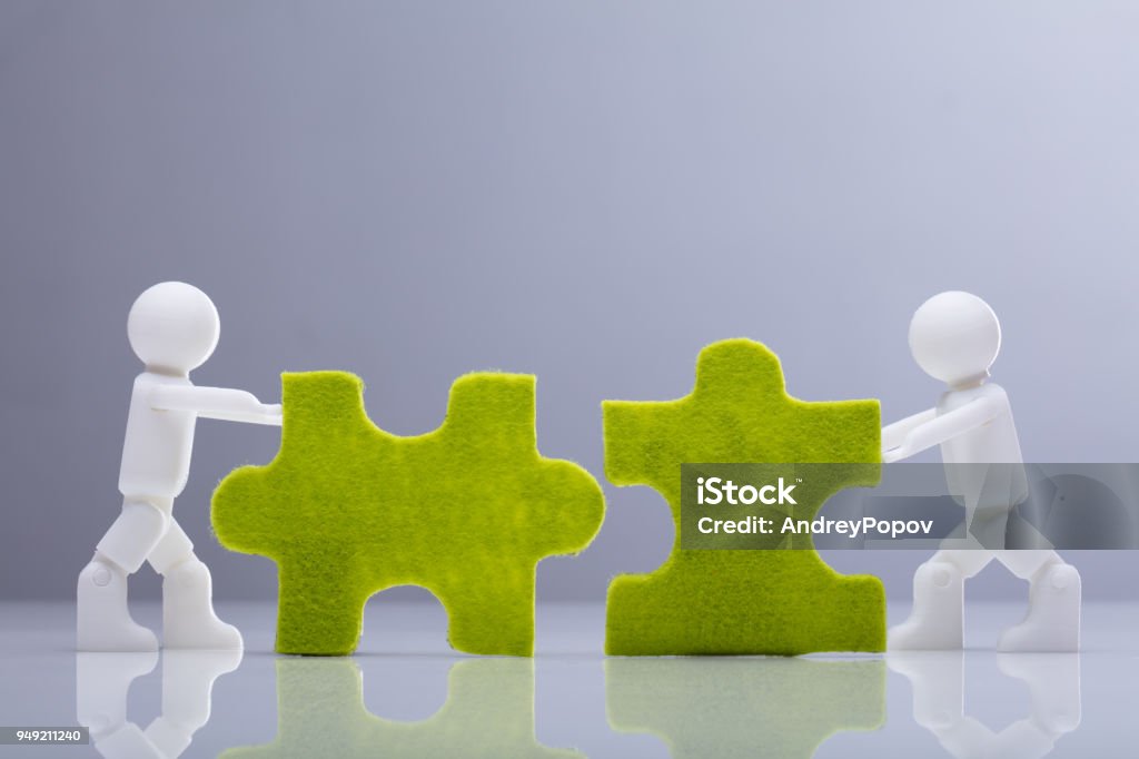 Two Miniature Human Figures Solving Green Jigsaw Puzzles Side View Of Two Miniature Human Figures Solving Green Jigsaw Puzzles On Grey Background Mergers and Acquisitions Stock Photo