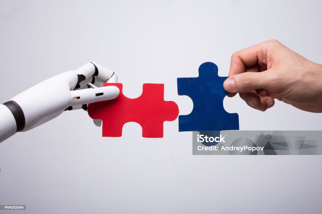 Robot And Human Hand Holding Jigsaw Puzzle Close-up Of Robot And Human Hand Holding Red And Blue Jigsaw Puzzle Artificial Intelligence Stock Photo