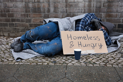 Homeless needy beggar male lying on cardboard box with sign HELP, asking for help. Wearing old dirty clothes