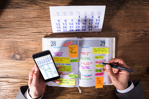 High Angle View Of A Businessperson With Mobile Phone Writing Schedule In Diary