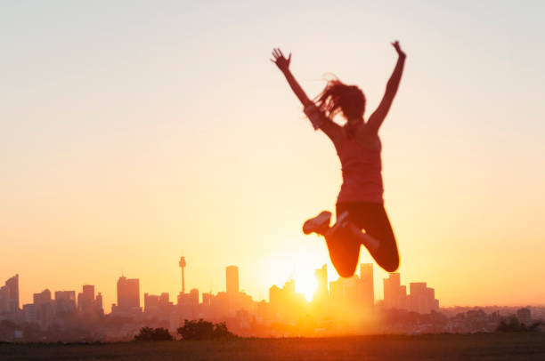 Sport women jumping and celebrating with arms raised. Sport women jumping and celebrating with arms raised. She is exercising at sunset or sunrise and is back lit. City of Sydney on the horizon in the background. Focus on background. She looks happy and has a sense of achievement. sydney sunset stock pictures, royalty-free photos & images