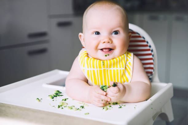 istock Baby's first solid food. Feeding. Messy smiling baby eating with a spoon in high chair. 949265224