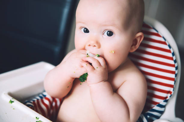 istock Feeding. Messy smiling baby eating with a spoon in high chair. Baby's first solid food. 949265486
