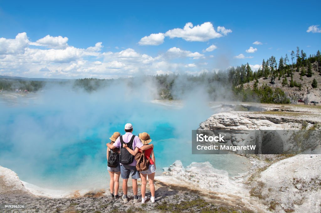 Family relaxing and enjoying beautiful view on vacation hiking trip. Family relaxing and enjoying beautiful view of gazer on vacation hiking trip. Father with arms around his family. Excelsior Geyser from the Midway Basin in Yellowstone National Park. Wyoming, USA Yellowstone National Park Stock Photo