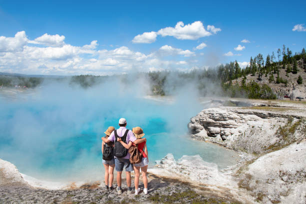Family relaxing and enjoying beautiful view on vacation hiking trip. Family relaxing and enjoying beautiful view of gazer on vacation hiking trip. Father with arms around his family. Excelsior Geyser from the Midway Basin in Yellowstone National Park. Wyoming, USA american tourism stock pictures, royalty-free photos & images