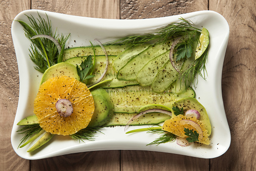 Close up of healthy salad with avocado, orange, cucumber and herbs on square white plate on natural wooden background. Top view.