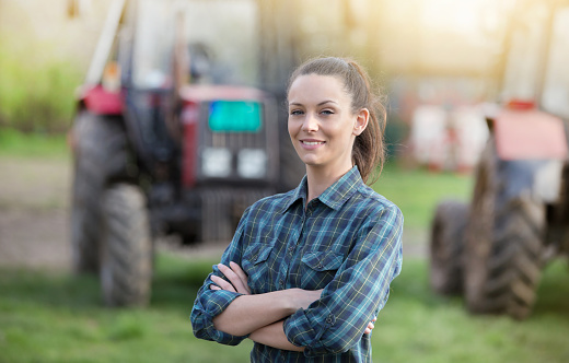 Young pretty farmer woman standing on farmland with crossed arms and tractors in background