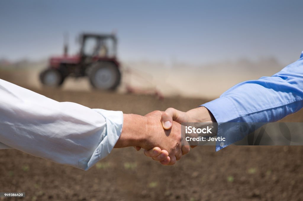 Farmers shaking hands in front of tractor in field Two businessmen shaking hands in field with tractor working in background. Agribusiness concept Agriculture Stock Photo