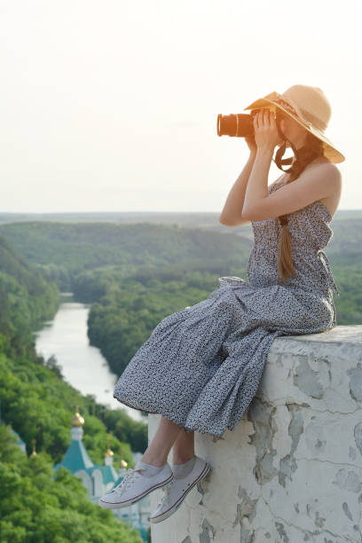 istock Girl sits on hill with camera. Forest, temple and river below 949394652