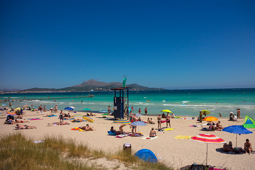 Alcudia, Mallorca, Spain - July 05, 2017: Sandy beach and clean blue sea panoramic image. Many people sunbathing. Lifeguard.