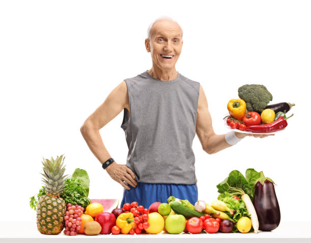 istock Senior in sportswear behind a table with fruit and vegetables holding a plate 949458526