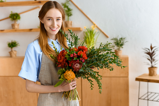 beautiful young florist holding bouquet and smiling at camera in flower shop