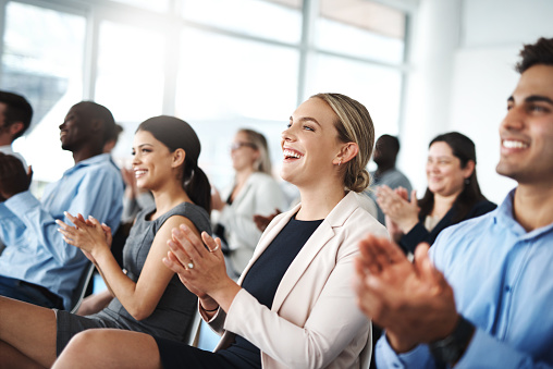 Happy young asian businesswoman clapping hands for presentation during a meeting in an office boardroom with colleagues. Diverse group of smiling businesspeople sitting in a row on a panel as audience and applauding after an inspiring and motivating talk