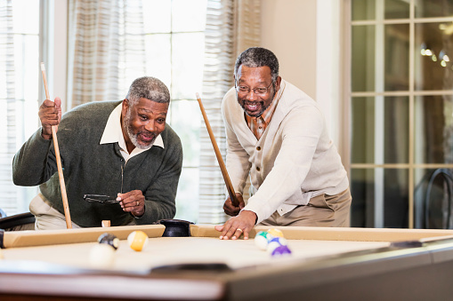 Two African-American men having fun shooting pool. The mature man, in his 50s, has just hit the cue ball with his cue stick.  He and his friend, a senior man in his 60s, are watching to see the results of his shot.
