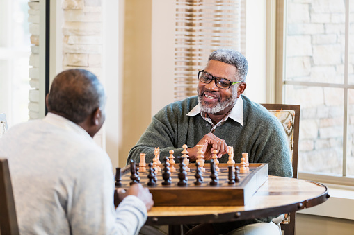 Two African-American men sitting at a chess board face to face, talking and smiling. The view is from over the shoulder of one man and the focus is on his friend, a senior man in his 60s wearing eyeglasses.