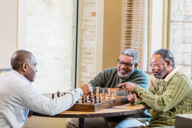 Three African-American men playing chess A group of three mature and senior African-American men in their 50s and 60s playing chess.  One man is moving a piece while his friends watch and discuss strategy. senior chess stock pictures, royalty-free photos & images