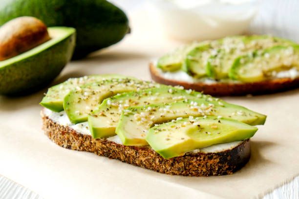 Vegan sandwich, rye bread toast, avocado slices, vegenaise sauce & raw arugula. Toasted sourdough, eggless mayonnaise, wooden table, whole & halved. National avocado day concept. Close up, background. Vegan sandwich, rye bread toast, avocado slices, vegenaise sauce & raw arugula. Toasted sourdough, eggless mayonnaise, wooden table, whole & halved. National avocado day concept. Close up, background. toasted bread stock pictures, royalty-free photos & images