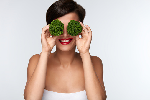Food For Health. Beautiful Smiling Woman With Red Lips Holding Organic Green Broccoli Covering Her Eyes. Portrait Of Happy Girl Playing With Vegetable. Healthy Diet Nutrition Concept. High Resolution