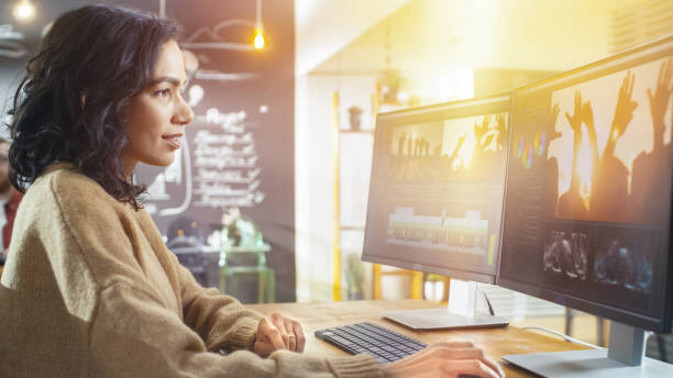 schöne junge frau kameramann arbeitet in der kreativagentur auf ihrem pc, bearbeiten von video-und audiomaterial. im hintergrund ihrer kollegen und tafel mit plänen. - loft apartment design studio office studio stock-fotos und bilder