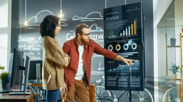 istock Female Developer and Male Statistician Use Interactive Whiteboard Presentation Screen to Look at Charts, Graphs and Growth Statistics. They Work in the Stylish Creative Office. 949561058