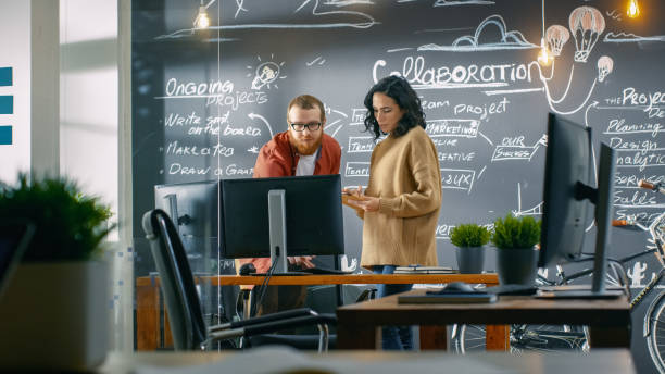 istock Female Designer Talks with Male Developer, They Look at Personal Computer Screen. In the Background Blackboard Wall with Project Plan. They Work in Creative Office. 949561154