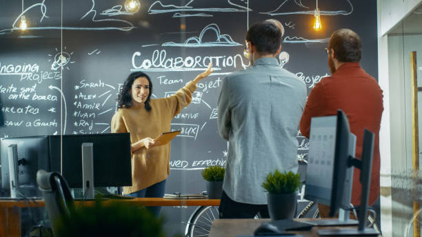 chef de projet femelle tient une réunion avec ses collègues de travail, elle détient ordinateur tablette et montre son dessin de plan d’un mur de blackboard. ils travaillent dans l’agence creative. - chef de projet photos et images de collection