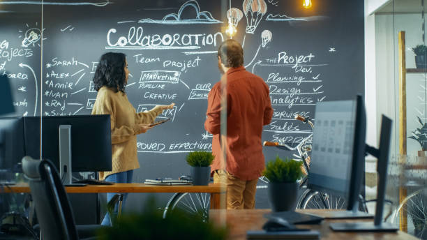 istock Female Project Manager Holds Tablet Computer and Shows Plan Drawn on a Blackboard Wall to Male Developer. They Work in the Creative Agency. 949561164