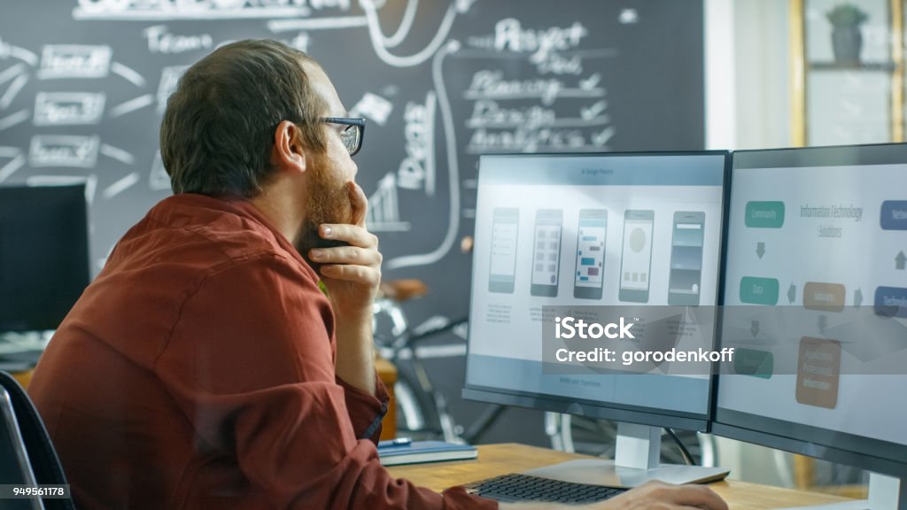 Male Mobile Video Game Application Developer Works on a Personal Computer, Designing Wireframe, He's Smooths the Beard in Thinking Gesture. He Works in the Creative Indie Studio. New Business Stock Photo