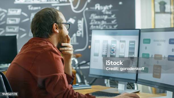 Male Mobile Video Game Application Developer Works On A Personal Computer Designing Wireframe Hes Smooths The Beard In Thinking Gesture He Works In The Creative Indie Studio Stock Photo - Download Image Now