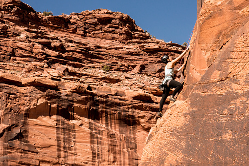 Female rock climber using climbing gear to climb up a sheer sandstone rock wall near Moab Utah