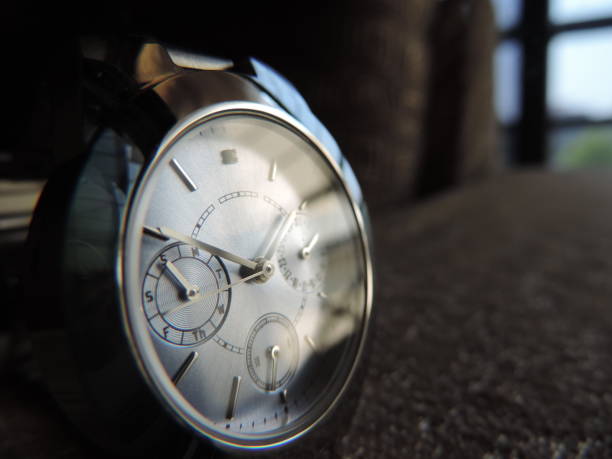metallic chrome wrist watch with day week and month hand placed near a window resting on a base with light reflected from its glass cover