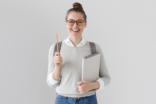 Studio shot of positive student girl isolated on grey background wearing glasses, sweater and jeans, backpack, pointing up with pencil, holding notebook, looking excited, full of energy and ideas.