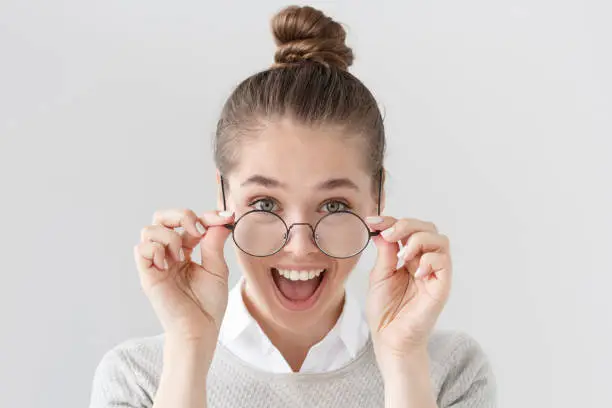 Photo of Indoor portrait of excited European female with hair tied in bun isolated on grey background with stylish black-rimmed eyeglasses, her mouth open wide in surprise with positive news or good offer.