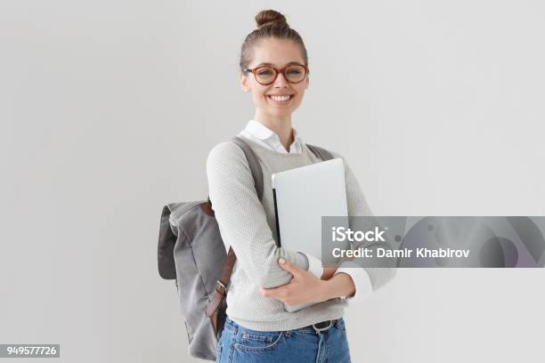 Foto de Foto Interior Da Faculdade Estudante Menina Isolada Em Um Fundo Cinza Sorrindo Para A Câmera Pressionando O Laptop Para Peito Usando Mochila Pronta Para Ir Para Estudos Iniciar Novo Projeto E Sugerir Novas Ideias e mais fotos de stock de Estudante