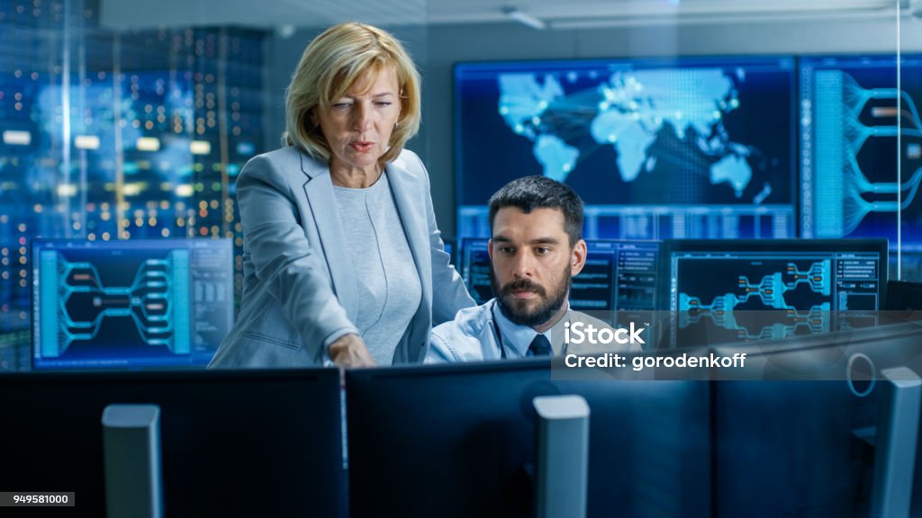In the System Monitoring Room Senior Supervisor Controls Work of the Operator. They're Surrounded by Monitors Showing Relevant Technical Data. Network Security Stock Photo