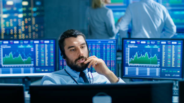 istock Stock Exchange Trader Makes a Deal with Big Client Over the Headset. He Works with Other Brokers and is Surround By Computers with Graphs and Ticker Numbers on Screens. 949581102
