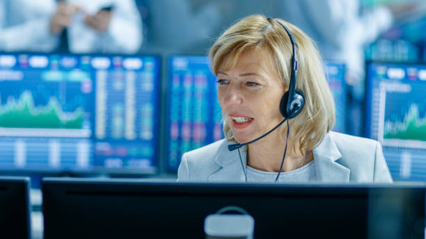 istock Chief Sales Force Representative Talks into the Headset. Behind Her People Working, Screens Show Stock Market Ticker Numbers and Graphs. 949581174
