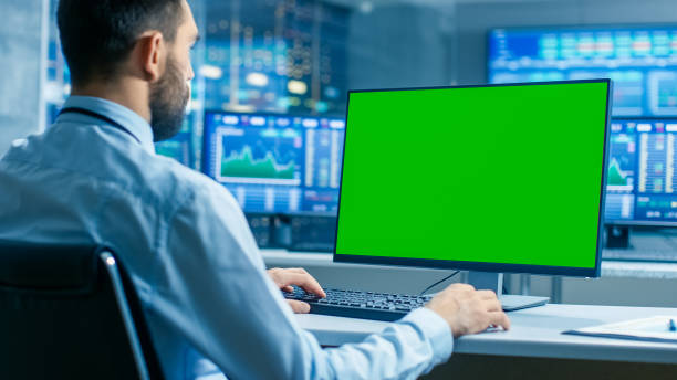 Stock Market Trader Working on a Computer with Isolated Mock-up Green Screen. In the Background Monitors Show Stock Ticker Numbers and Graphs. Stock Market Trader Working on a Computer with Isolated Mock-up Green Screen. In the Background Monitors Show Stock Ticker Numbers and Graphs. looking over shoulder stock pictures, royalty-free photos & images