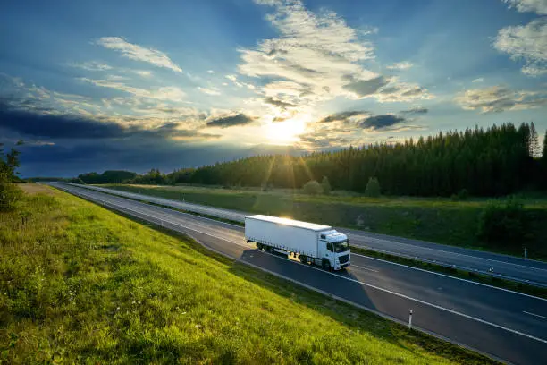 Photo of White truck driving on the highway in the countryside in the rays of the sunset with dramatic clouds