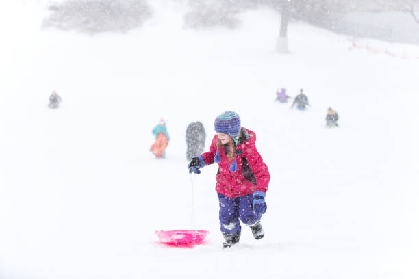 Young girl pulling her bright pink sled up a sledding hill after sledding down. Other children can be seen in the distance near the bottom of the hill. Taken on a snowy late winter/early springtime day on the bank of the Mississippi River.