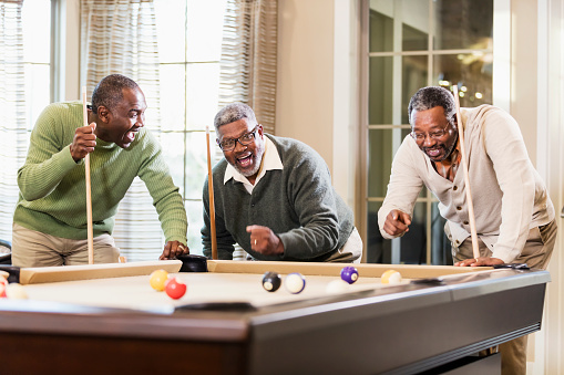 Three mature and senior African-American men in their 50s and 60s having fun shooting pool. They are excited about the last shot taken. The man in the middle is a senior man in his 60s. His friends are in their 50s.